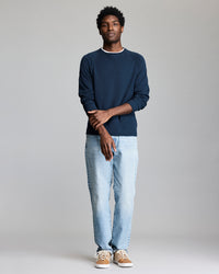 A person stands against a plain light gray background, wearing a navy blue long-sleeve shirt, light blue jeans, and tan sneakers with white laces. They stand with arms crossed and a relaxed expression.