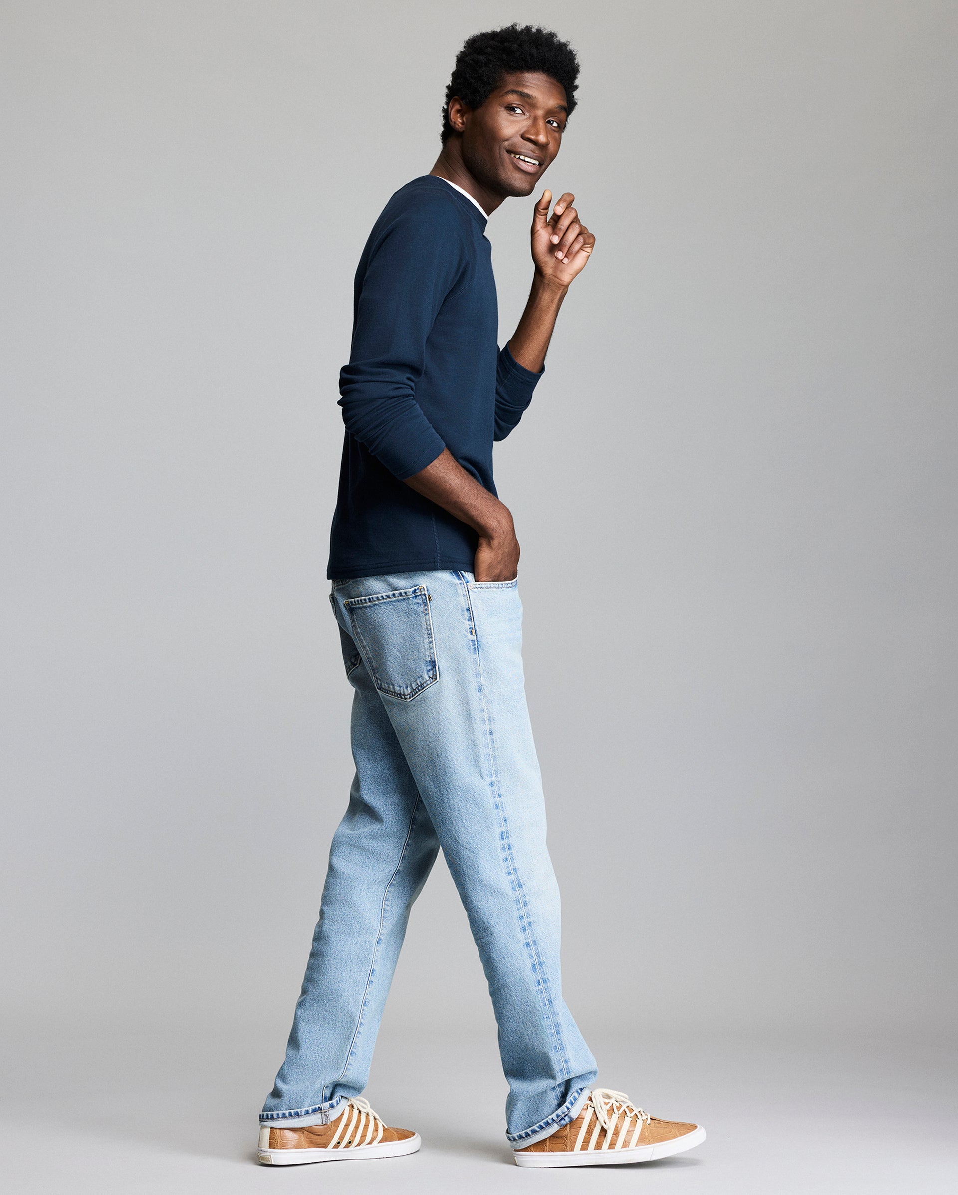 A man stands sideways, smiling and looking over his shoulder. He wears a navy long-sleeve shirt, light blue jeans, and brown sneakers with white stripes, against a plain light gray background.