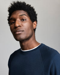A young man with short, curly hair wearing a navy blue sweatshirt over a white shirt looks confidently at the camera against a plain, light gray background.
