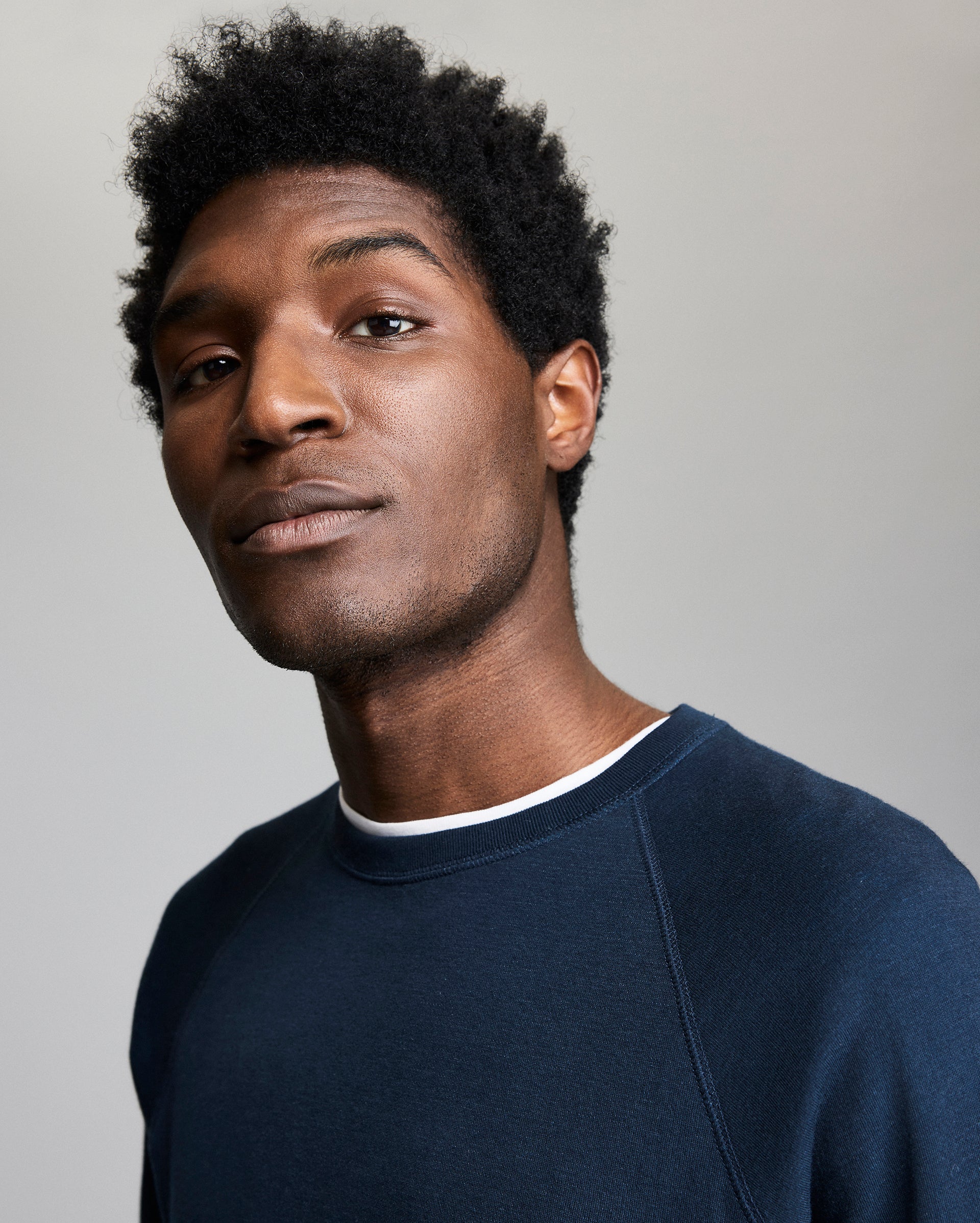 A young man with short, curly hair wearing a navy blue sweatshirt over a white shirt looks confidently at the camera against a plain, light gray background.