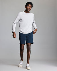 A man wearing a long-sleeve white shirt, dark blue shorts, and white sneakers stands against a plain gray background, looking forward with one hand in his pocket.
