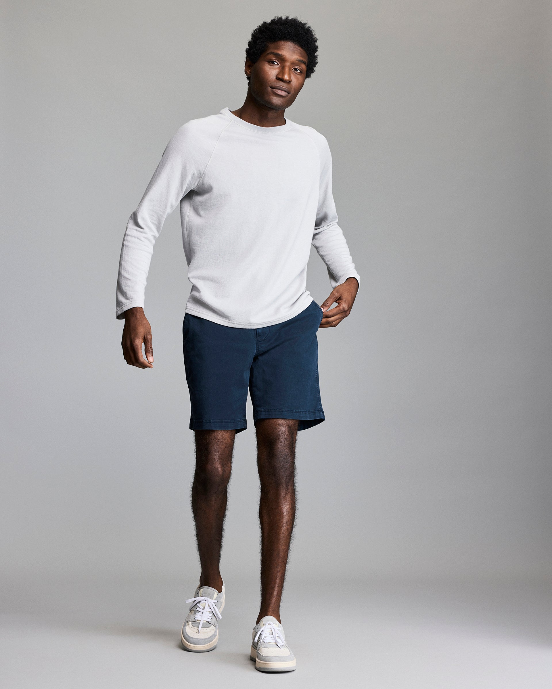 A man wearing a long-sleeve white shirt, dark blue shorts, and white sneakers stands against a plain gray background, looking forward with one hand in his pocket.