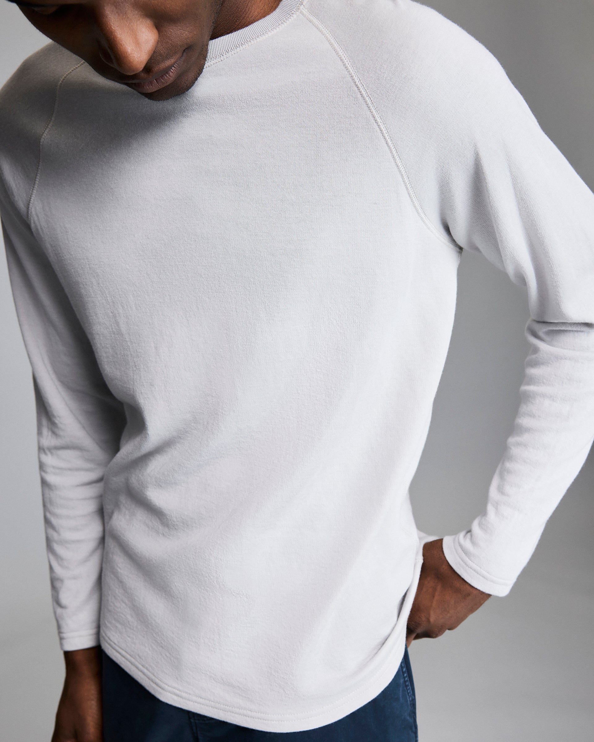 A person wearing a light gray, long-sleeve shirt and dark pants stands against a plain, neutral background, with their head turned downward and one hand resting on their hip.