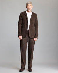 A man with short gray hair and beard wears a brown suit with a white t-shirt and brown shoes, standing against a plain light gray background.