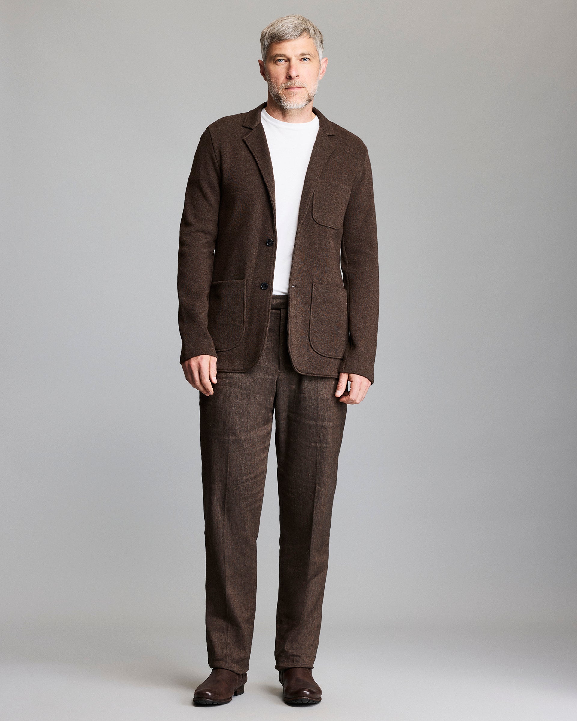A man with short gray hair and beard wears a brown suit with a white t-shirt and brown shoes, standing against a plain light gray background.
