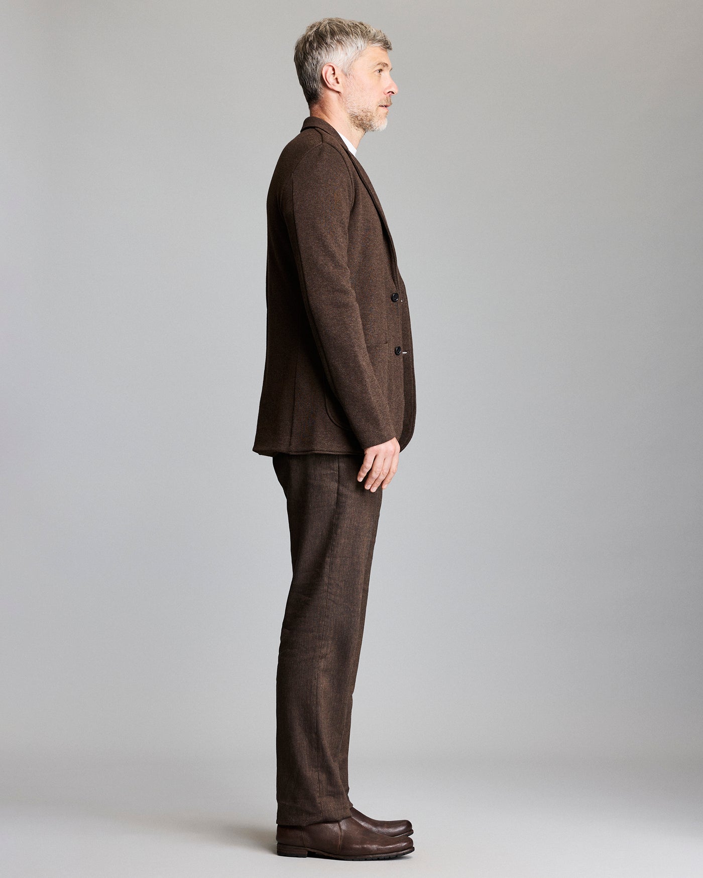 A man with short gray hair stands in profile against a plain background, wearing a brown suit and matching shoes.