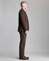A man with short gray hair stands in profile against a plain background, wearing a brown suit and matching shoes.