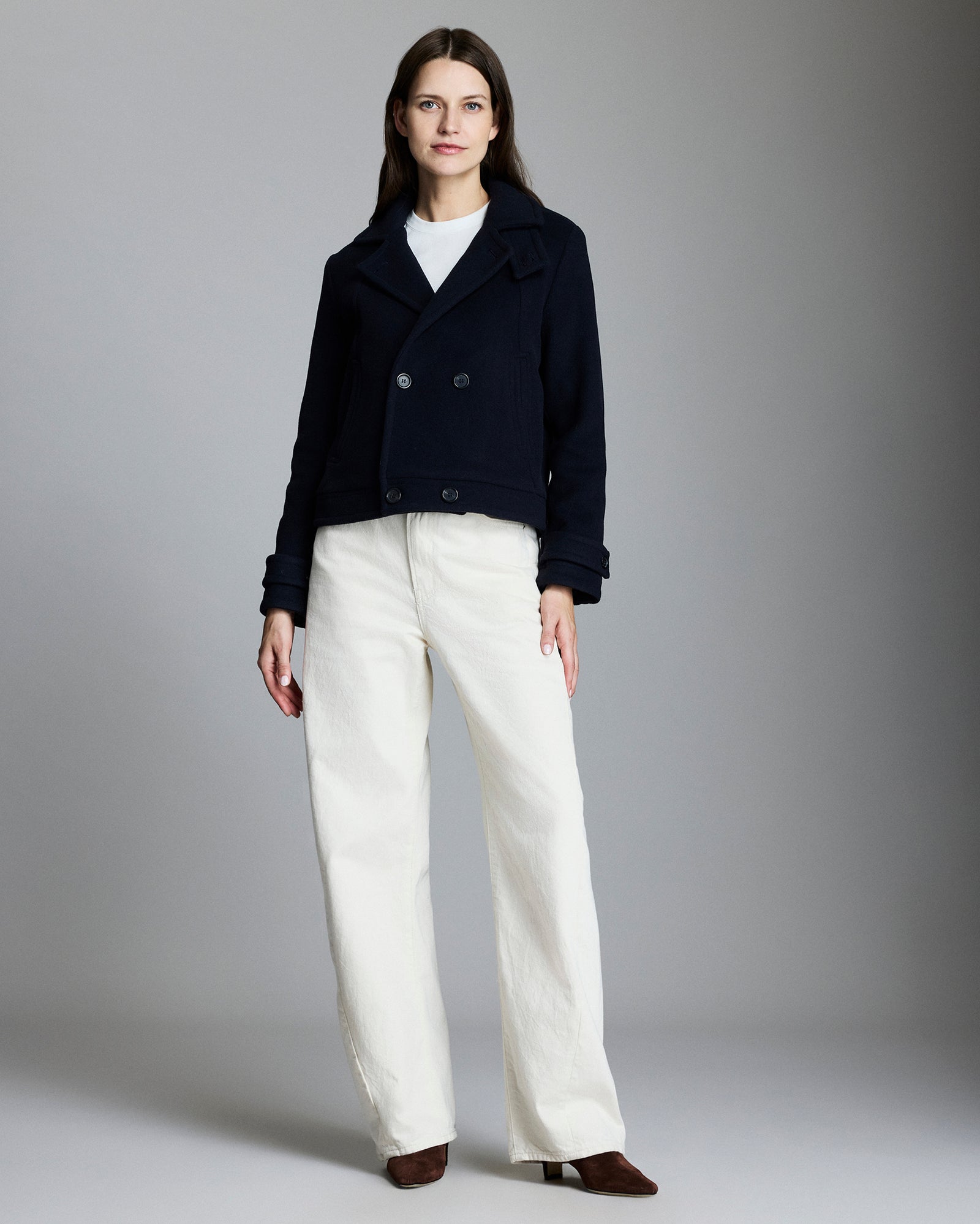 Woman wearing a dark coat and white pants against a gray background