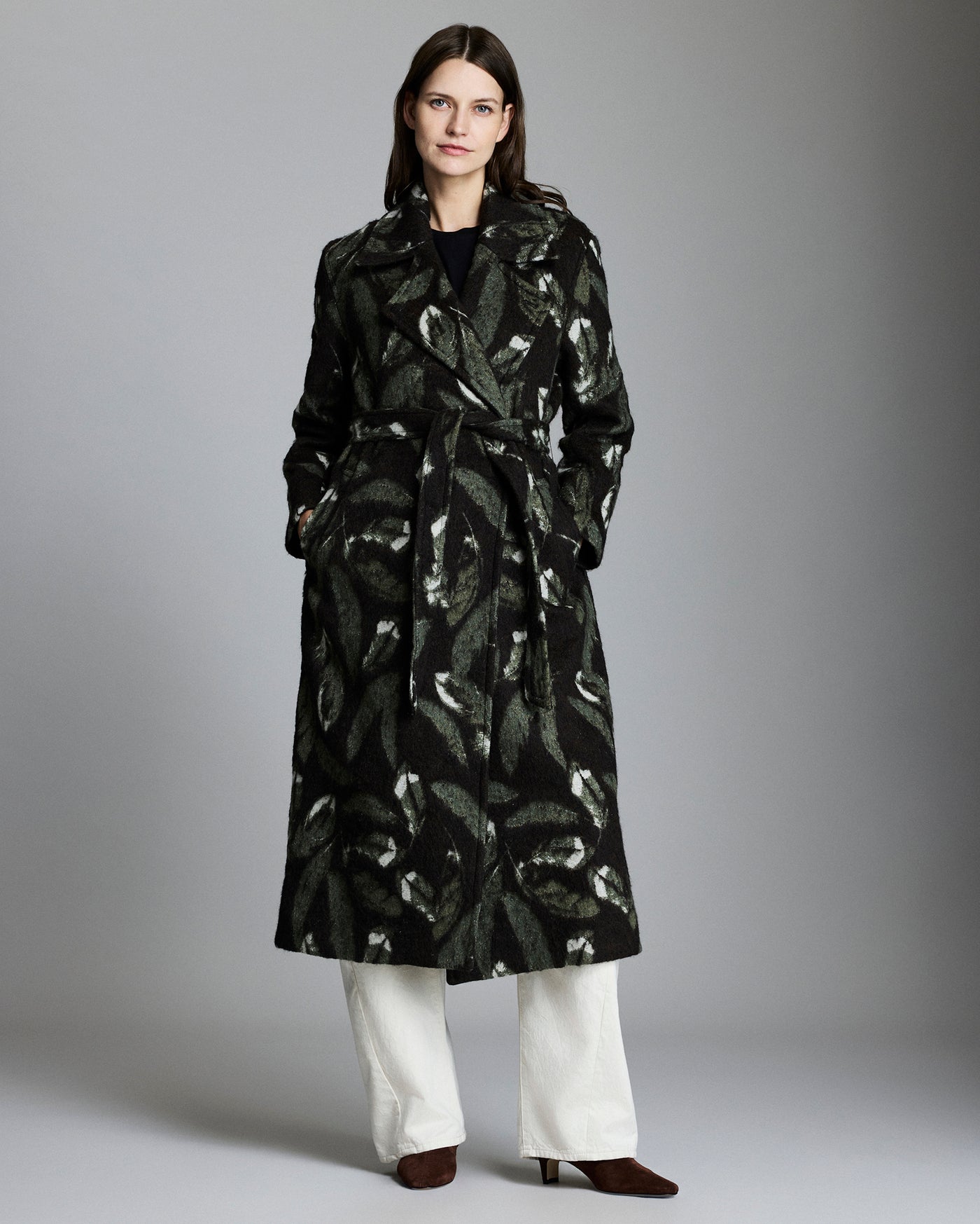 Woman wearing a black patterned coat with white pants on a gray background