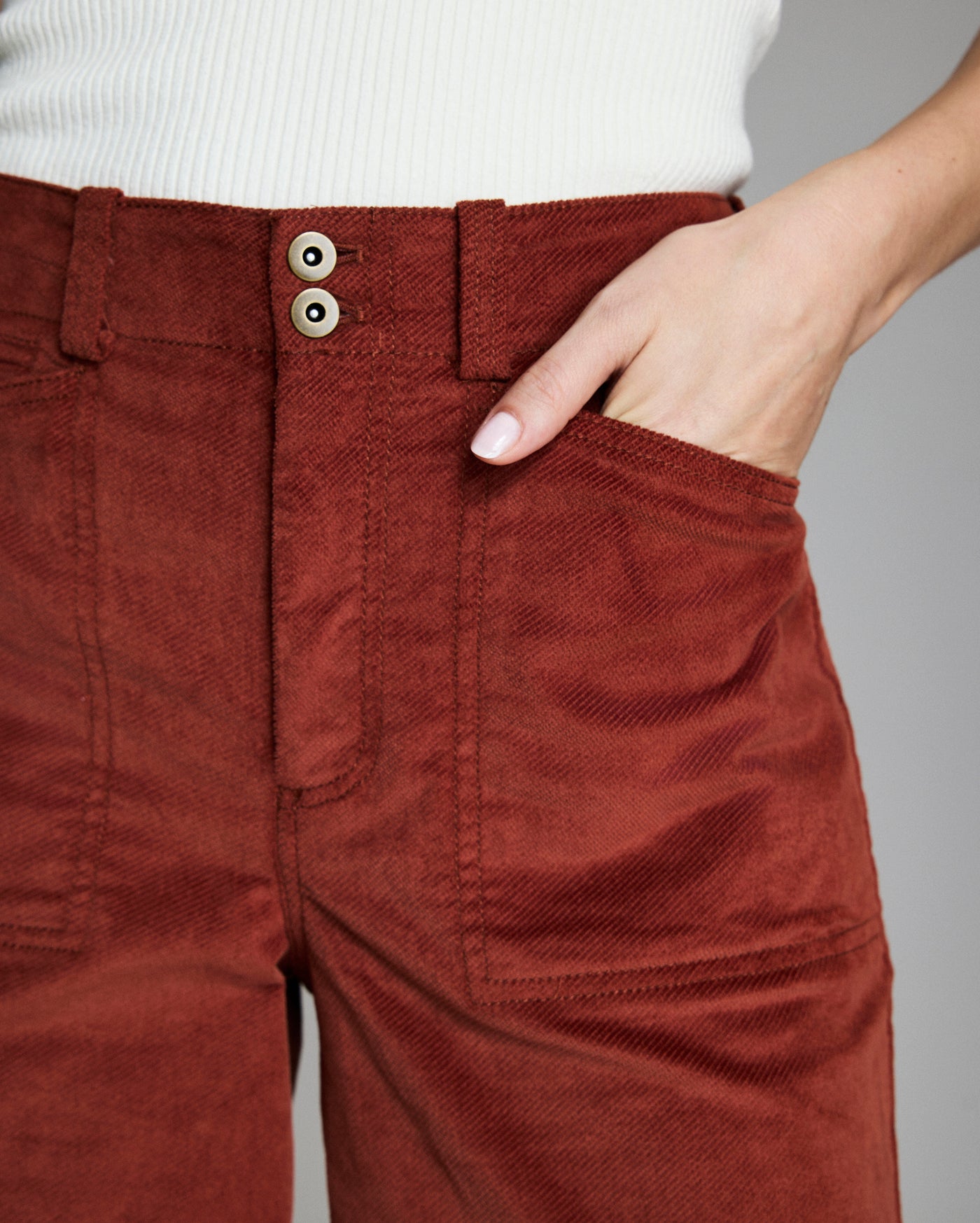 A person wearing high-waisted, rust-colored corduroy pants with a white ribbed top, with one hand placed in the pants pocket.