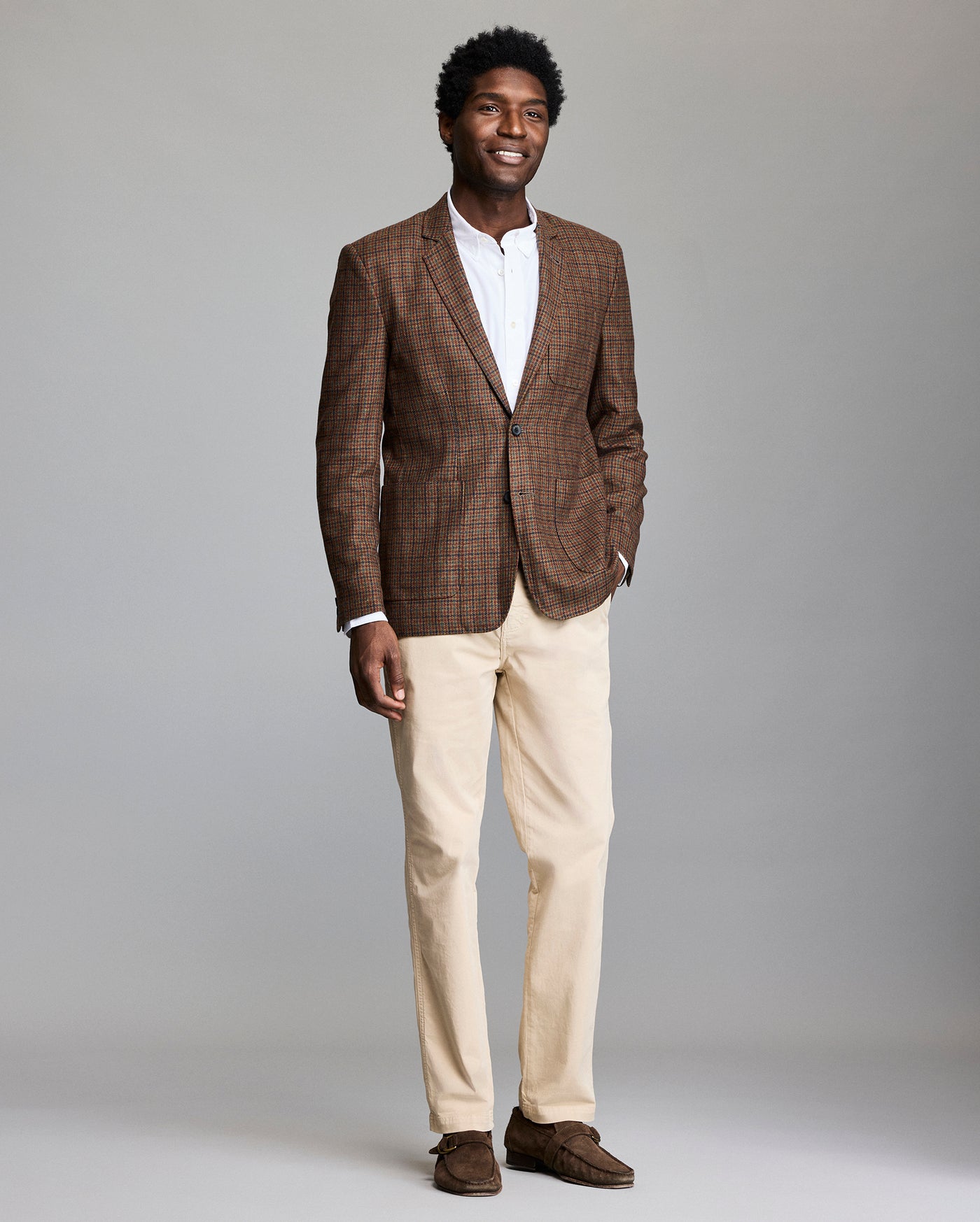 A man stands smiling against a plain background, wearing a brown plaid blazer, white dress shirt, beige pants, and brown loafers.