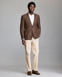 A man stands smiling against a plain background, wearing a brown plaid blazer, white dress shirt, beige pants, and brown loafers.
