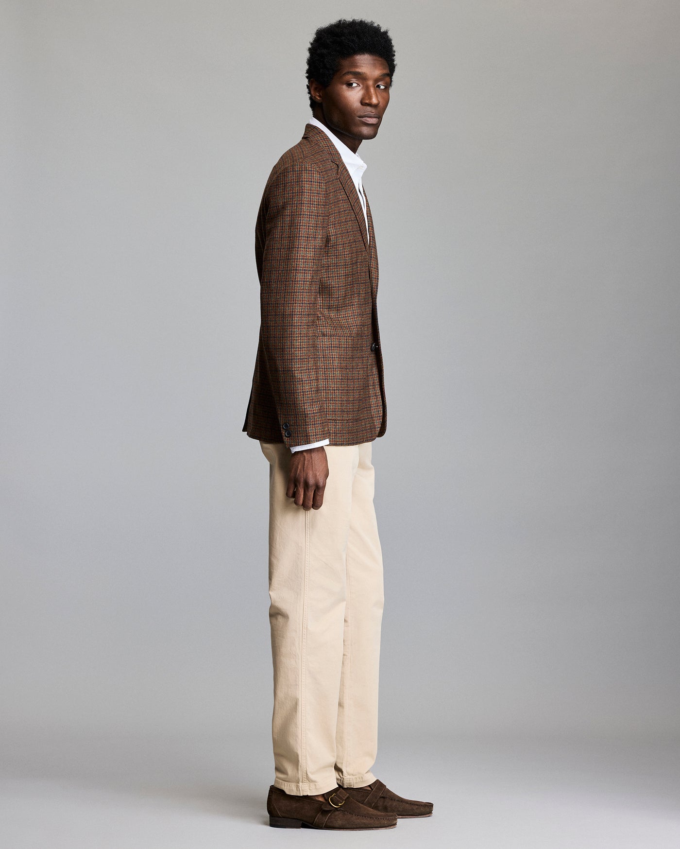 A man stands in profile against a plain background, wearing a brown plaid blazer, white shirt, beige trousers, and brown suede shoes.