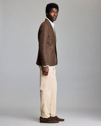 A man stands in profile against a plain background, wearing a brown plaid blazer, white shirt, beige trousers, and brown suede shoes.