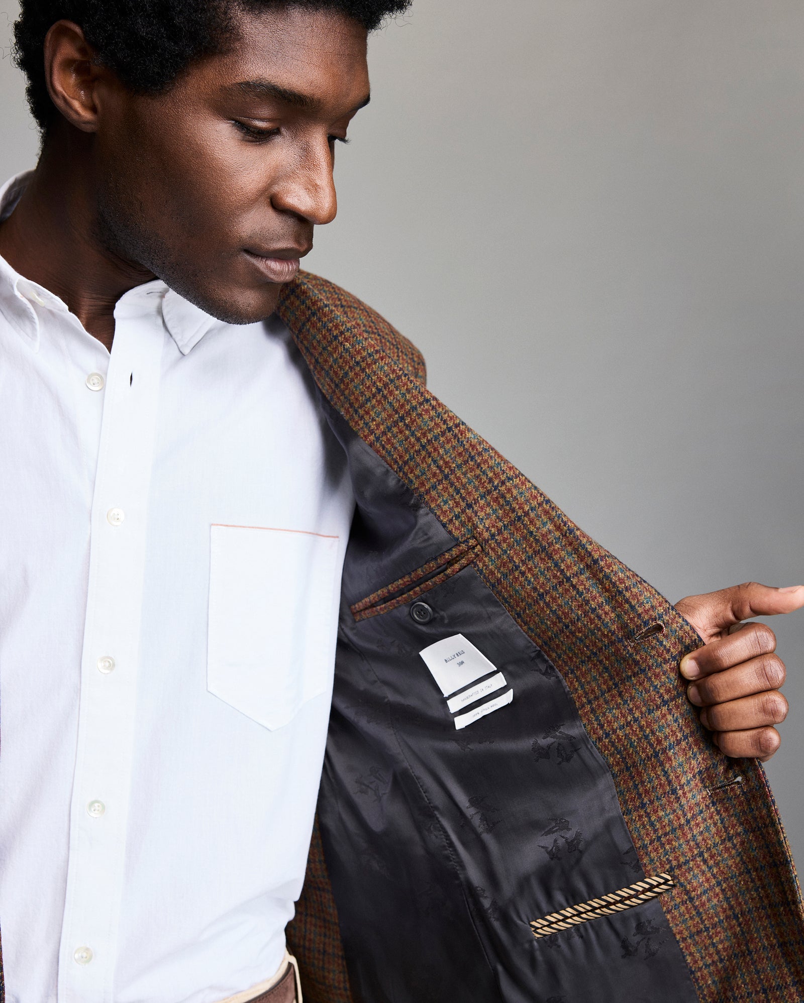 A person in a white button-up shirt holds open a brown plaid blazer, revealing the patterned black lining, a striped label, a pocket, and a gold chain detail inside the jacket.