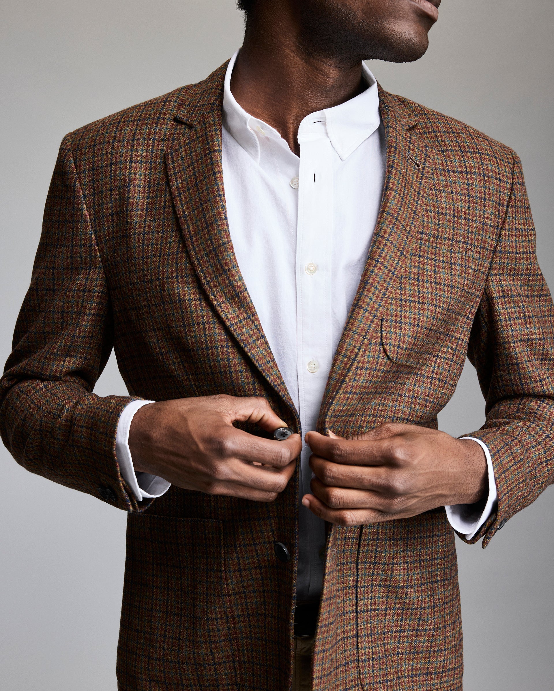 A man wearing a brown plaid blazer over a white dress shirt buttons his jacket, facing slightly to the side against a neutral background.