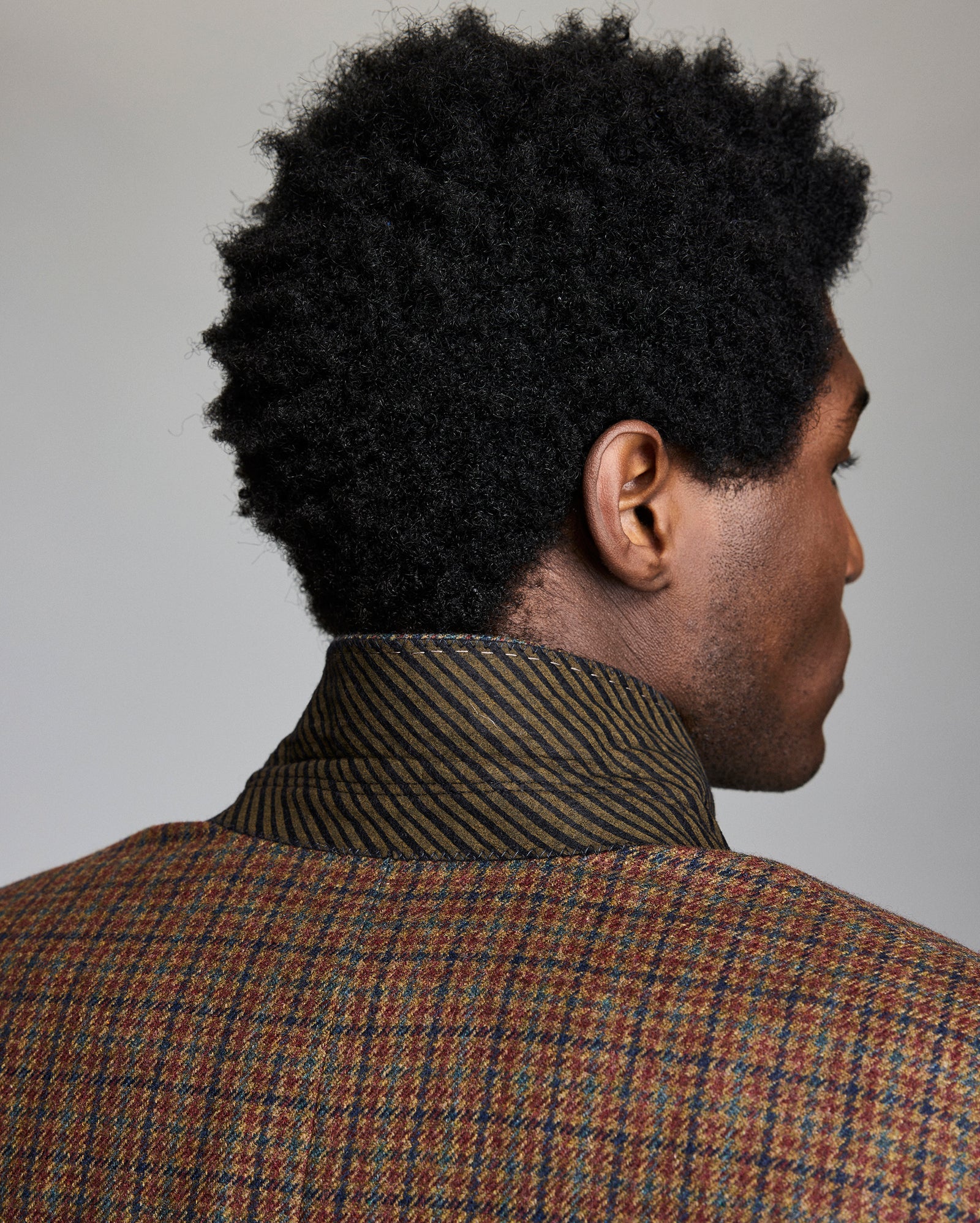 A man with short, curly black hair is shown from behind, wearing a brown plaid jacket with the collar flipped up, revealing a dark, diagonally-striped inner lining. The background is plain and neutral.