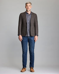 Man wearing a brown checkered blazer, plaid shirt, blue jeans, and brown shoes on a gray background