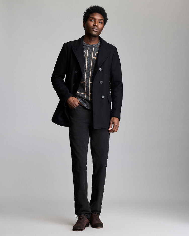 A man stands against a plain background wearing a black double-breasted coat, a patterned sweater, dark pants, and brown shoes. He has short curly hair and a neutral expression, with one hand in his pocket.