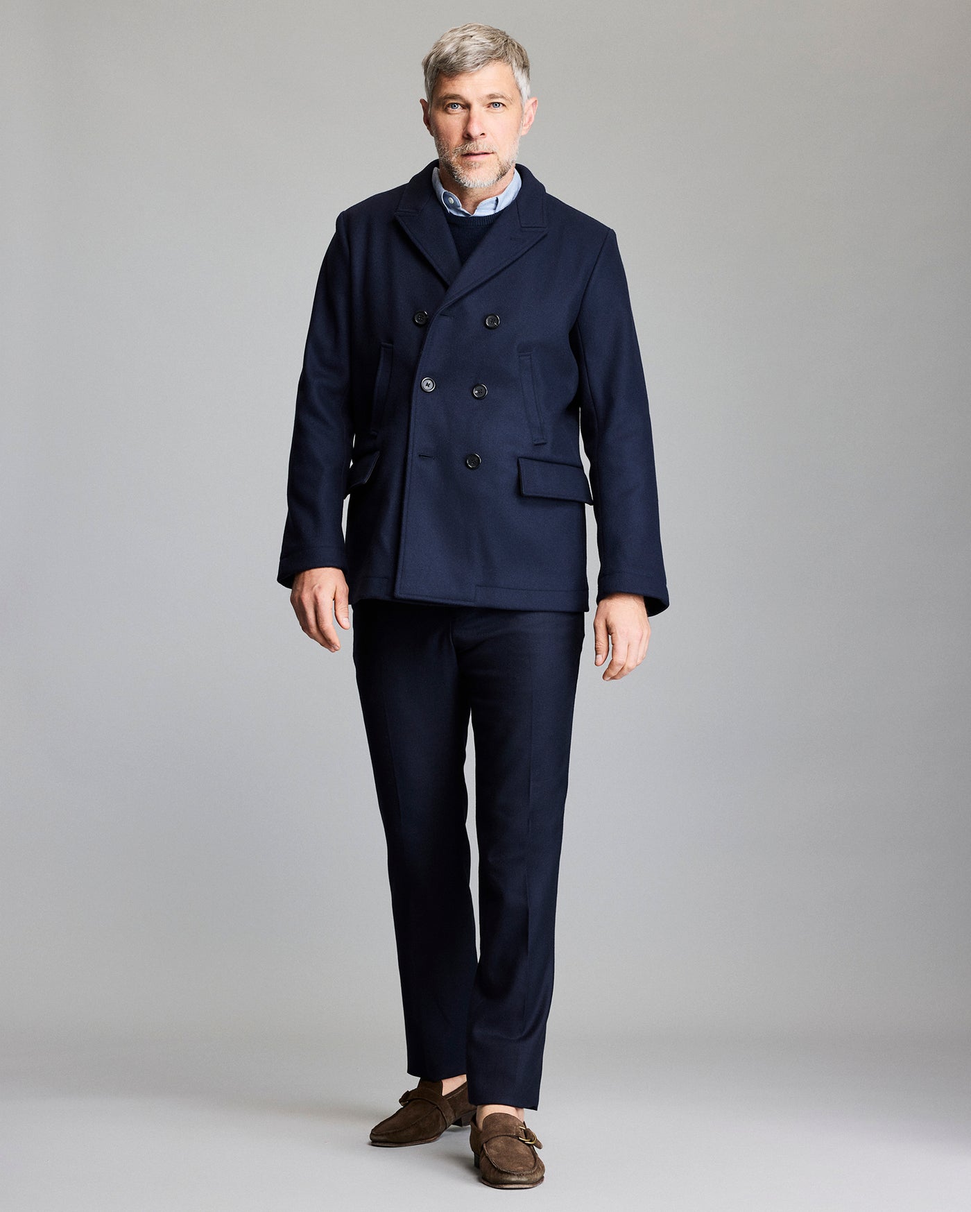Man wearing a navy peacoat and dark pants on a gray background