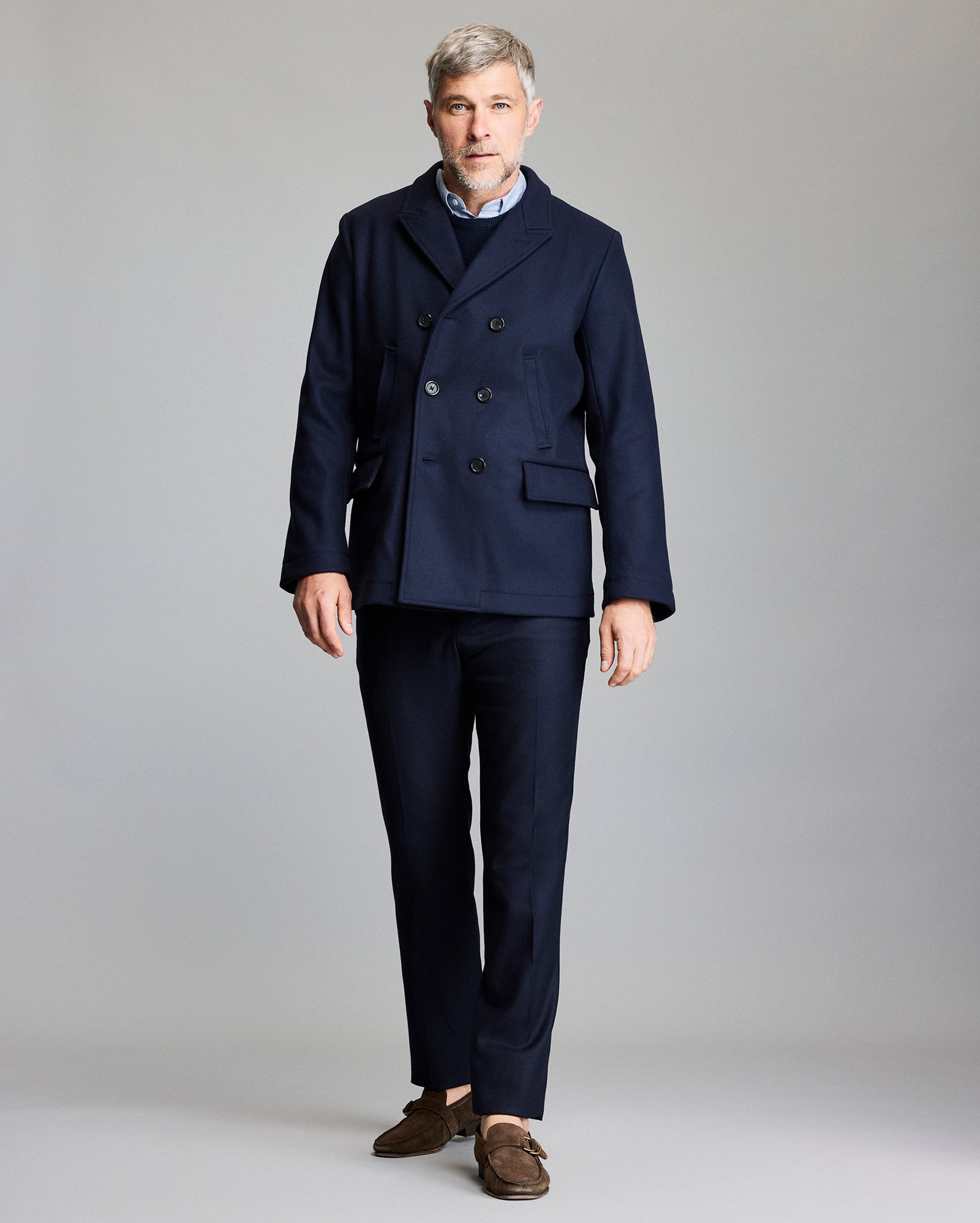 Man wearing a navy peacoat and dark pants on a gray background