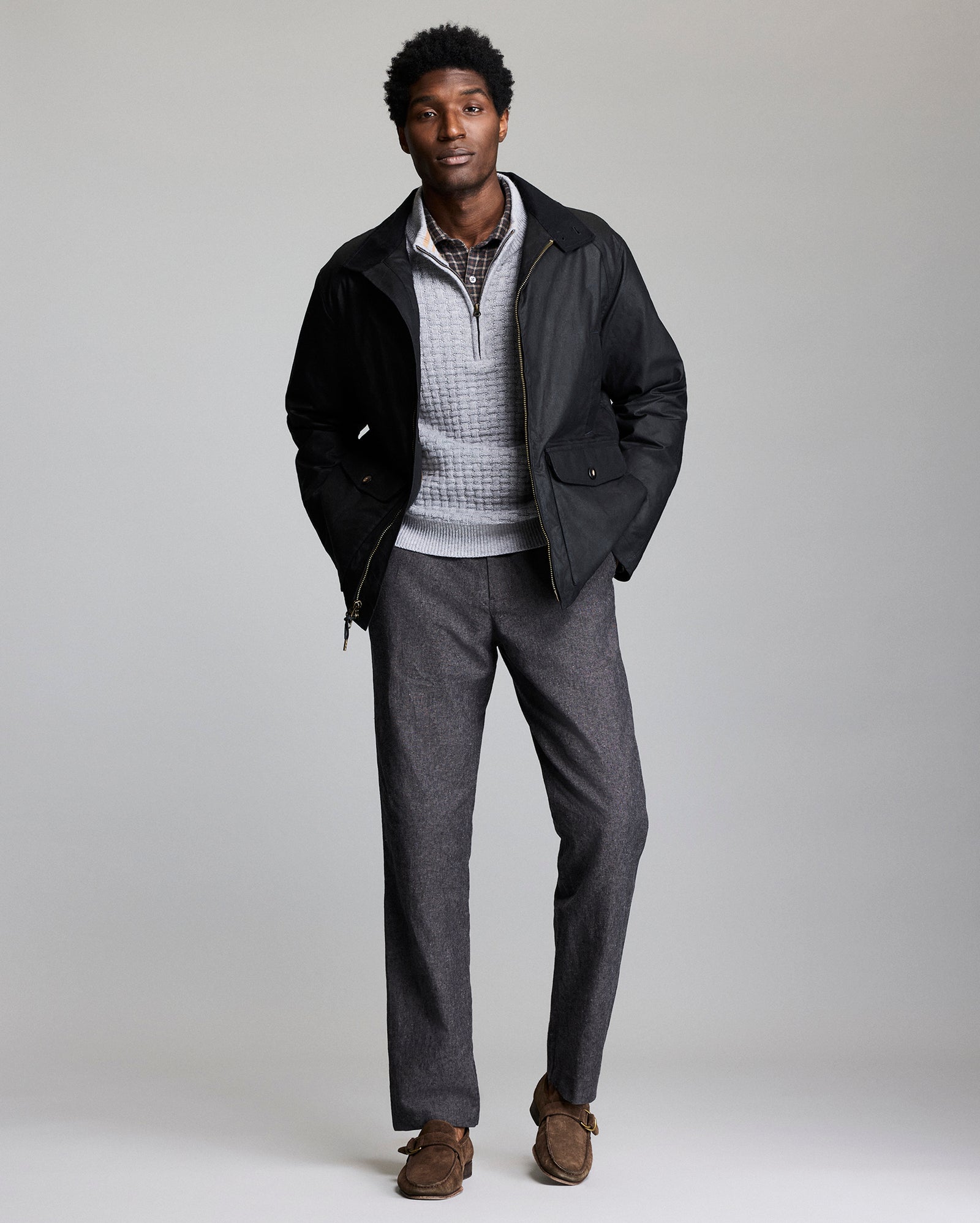 A man stands against a plain background wearing a black jacket over a light gray quarter-zip sweater, a checked shirt, gray trousers, and brown loafers. His hands are in his jacket pockets and he is looking at the camera.