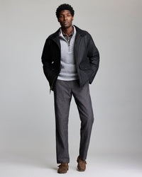 A man stands against a plain background wearing a black jacket over a light gray quarter-zip sweater, a checked shirt, gray trousers, and brown loafers. His hands are in his jacket pockets and he is looking at the camera.