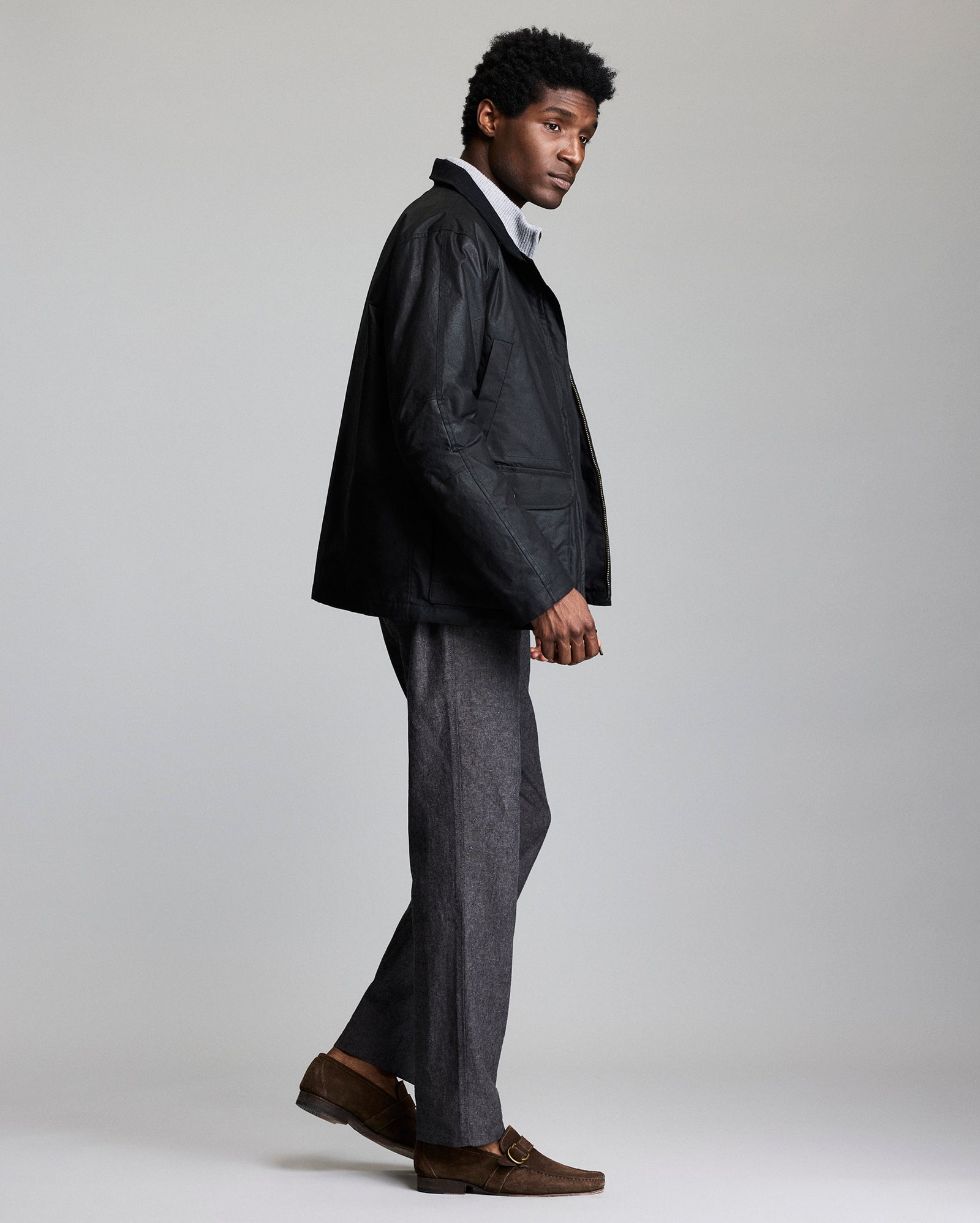A man stands sideways, wearing a black jacket over a white turtleneck, gray trousers, and brown loafers. He faces left against a plain light gray background.
