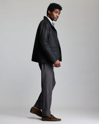 A man stands sideways, wearing a black jacket over a white turtleneck, gray trousers, and brown loafers. He faces left against a plain light gray background.