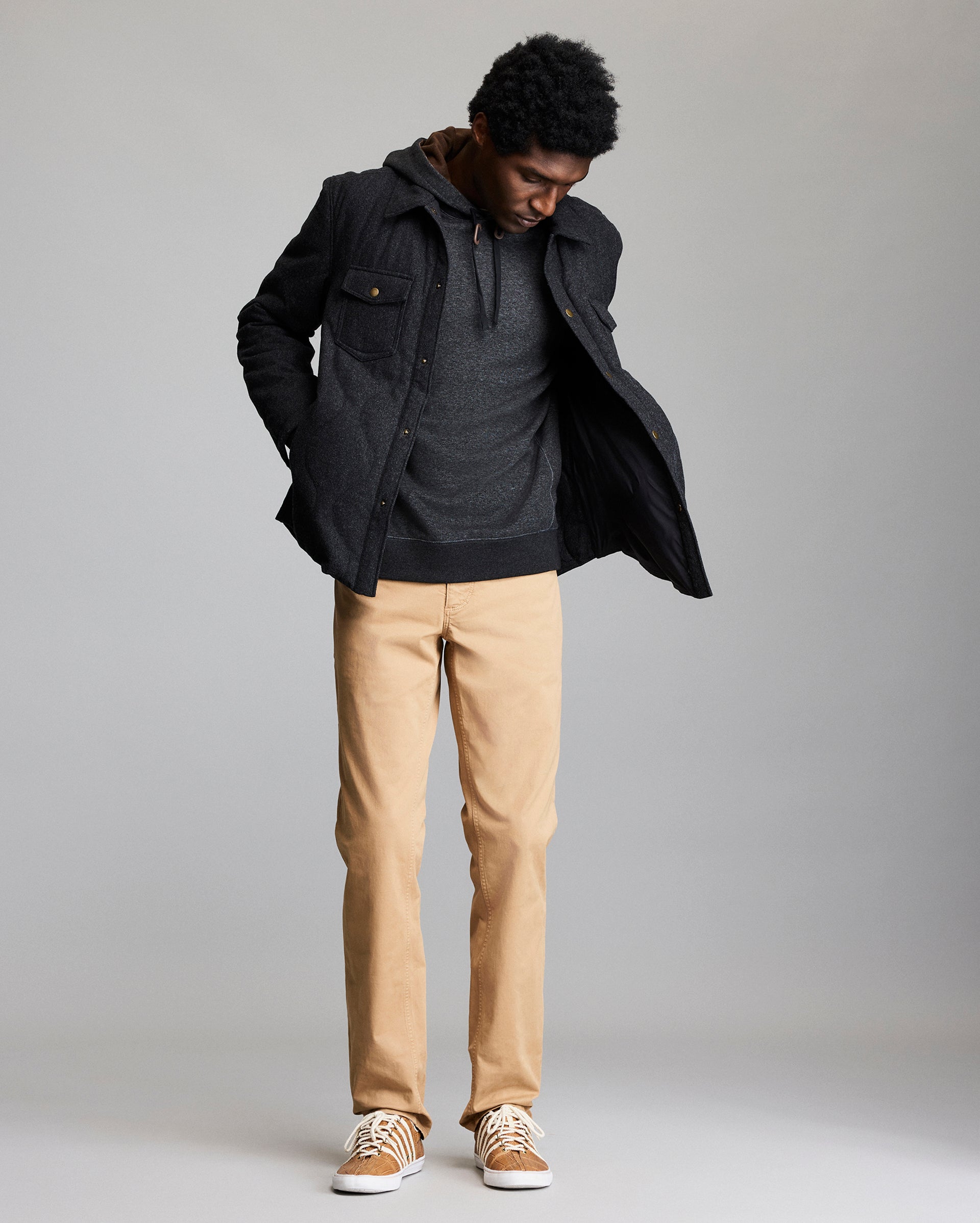 A man stands against a plain background, wearing a dark jacket over a gray hoodie, tan pants, and tan sneakers with white laces, looking down with his hands in his jacket pockets.