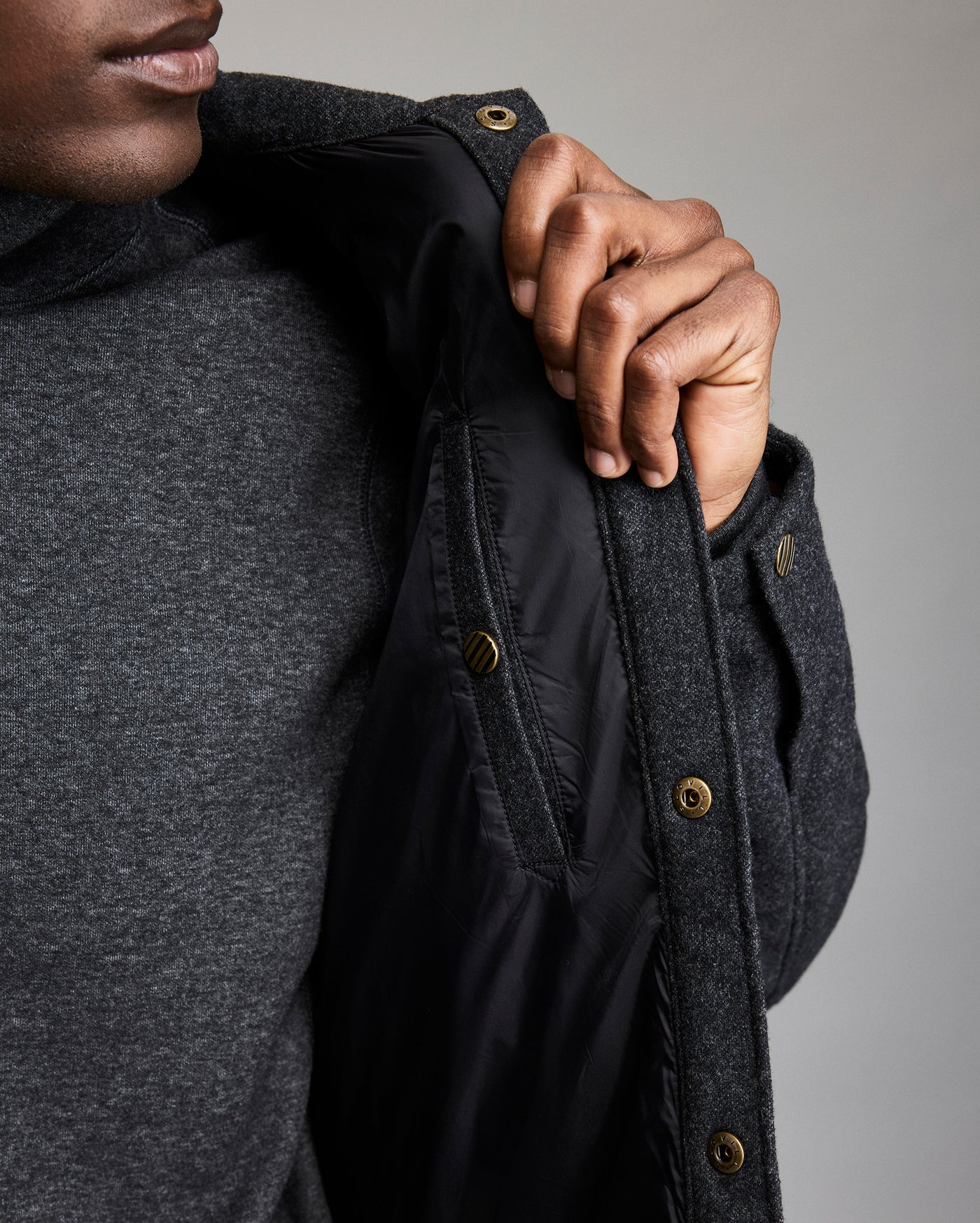 A person wearing a dark gray coat and sweater holds open the coat to reveal its black inner lining and zippered pocket. Only the lower half of the face and torso are visible.