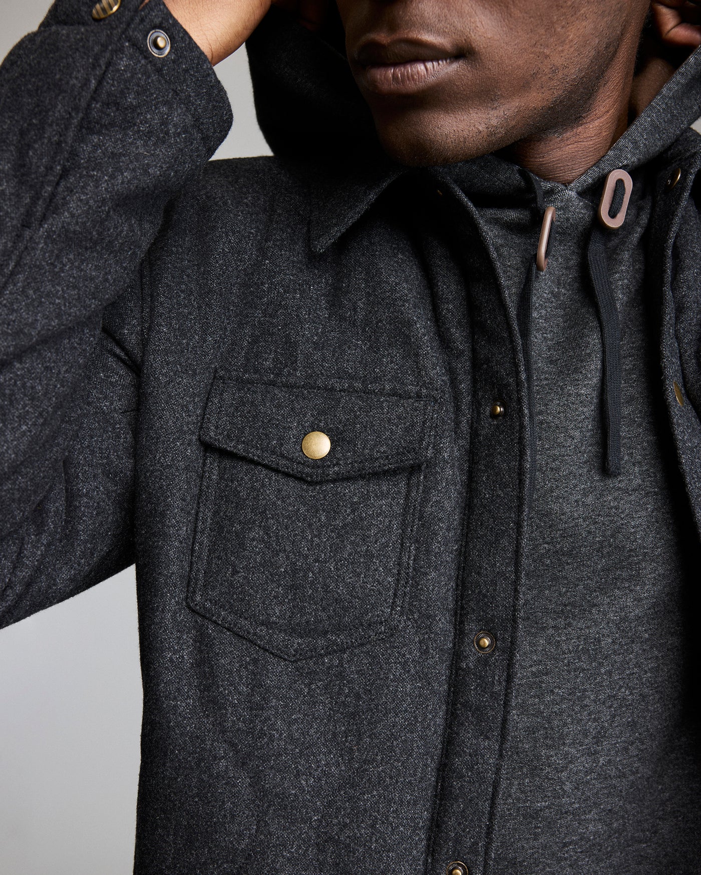 A close-up of a person wearing a dark gray button-up jacket with a chest pocket and a hoodie underneath. The persons face is partially out of frame, and they have a hand raised near their head.