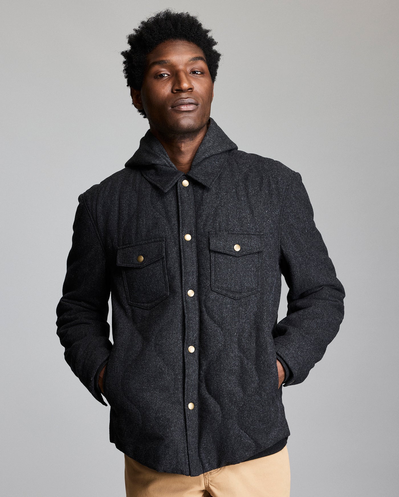 A man stands against a plain background wearing a dark, button-up jacket with flap pockets and tan pants. His hands are in his jacket pockets, and he looks confidently at the camera.
