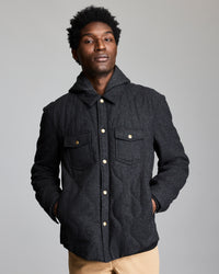 A man stands against a plain background wearing a dark, button-up jacket with flap pockets and tan pants. His hands are in his jacket pockets, and he looks confidently at the camera.