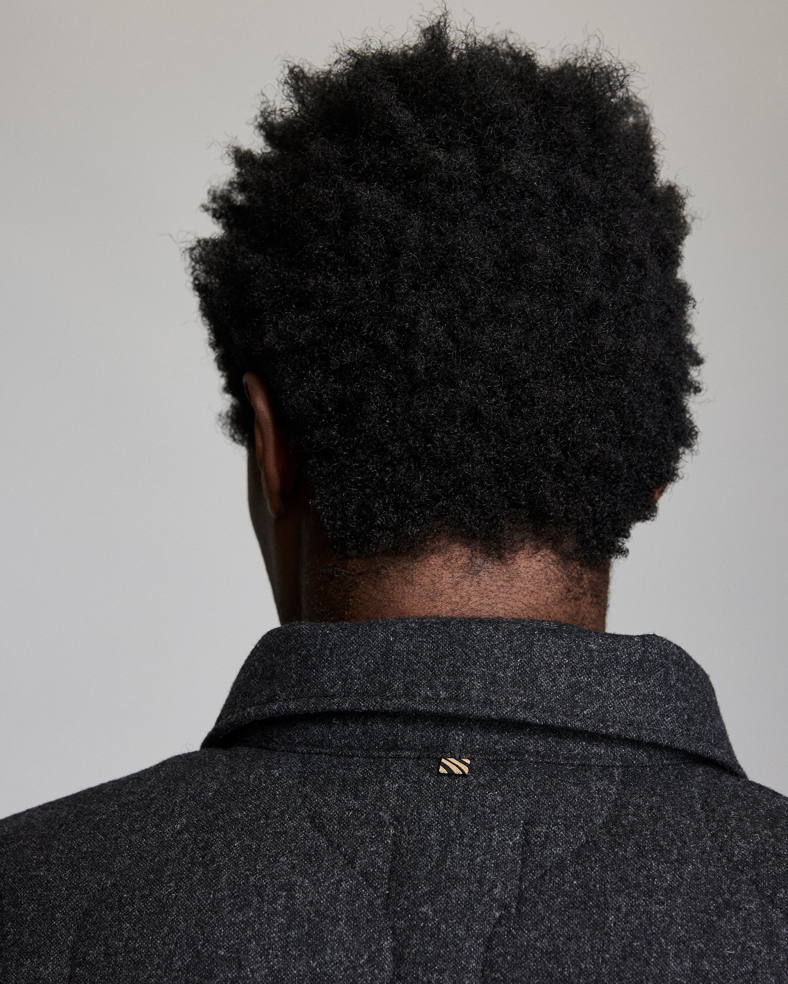 A person with short, curly black hair is shown from behind, wearing a dark textured coat with a small gold pin on the collar. The background is plain and light-colored.