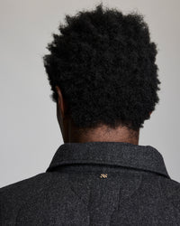 A person with short, curly black hair is shown from behind, wearing a dark textured coat with a small gold pin on the collar. The background is plain and light-colored.