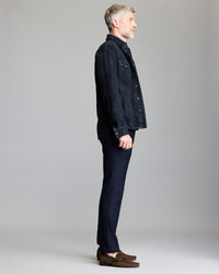 A man with short gray hair stands in profile against a plain background, wearing a dark jacket, dark pants, and brown loafers.