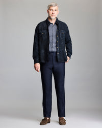 A man with gray hair and a short beard stands facing forward, wearing a dark jacket over a patterned shirt, dark trousers, and brown loafers, against a plain light gray background.