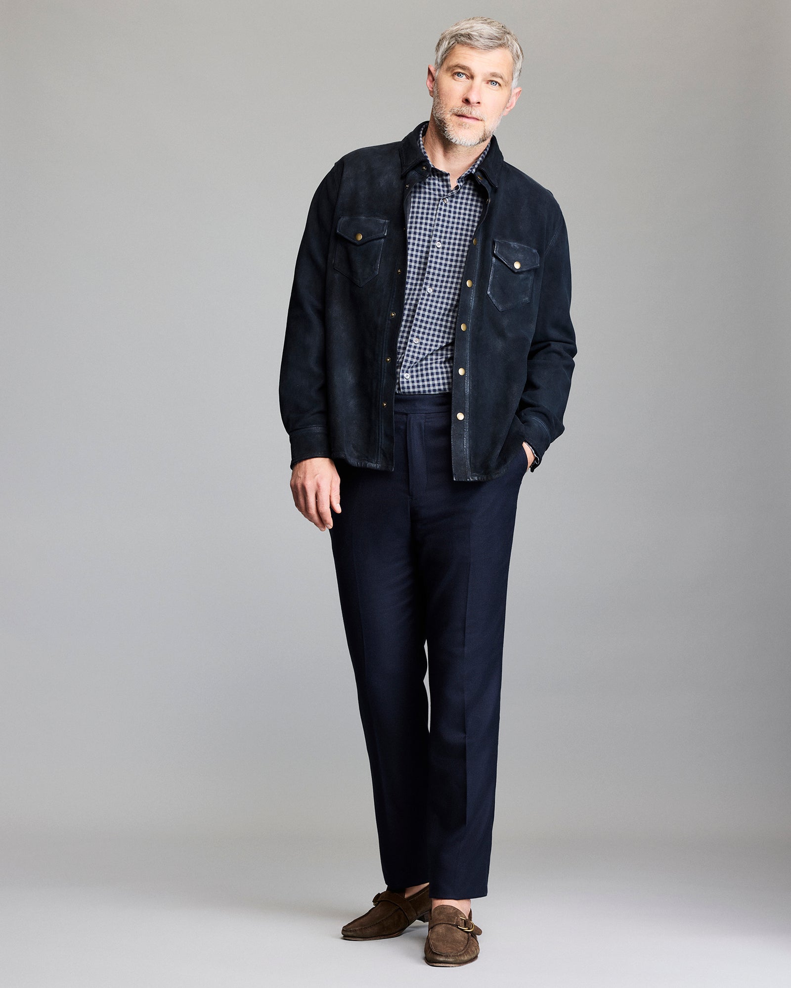 A man with gray hair and a beard stands against a plain background, wearing a dark suede jacket over a checkered shirt, navy trousers, and brown loafers. He has one hand in his pocket and a neutral expression.