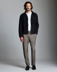 A man with wavy hair stands against a plain background, wearing a black jacket over a white shirt, brown checked trousers, and black shoes. One hand is in his pocket and he looks directly at the camera.