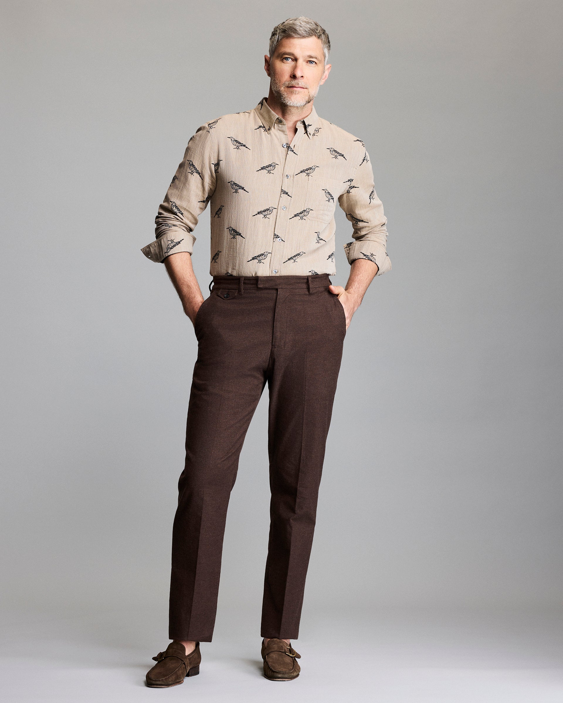 A man with gray hair and a beard stands against a plain background, wearing a beige button-up shirt with a bird print, dark brown dress pants, and brown loafers. His hands are in his pockets and he looks confidently ahead.
