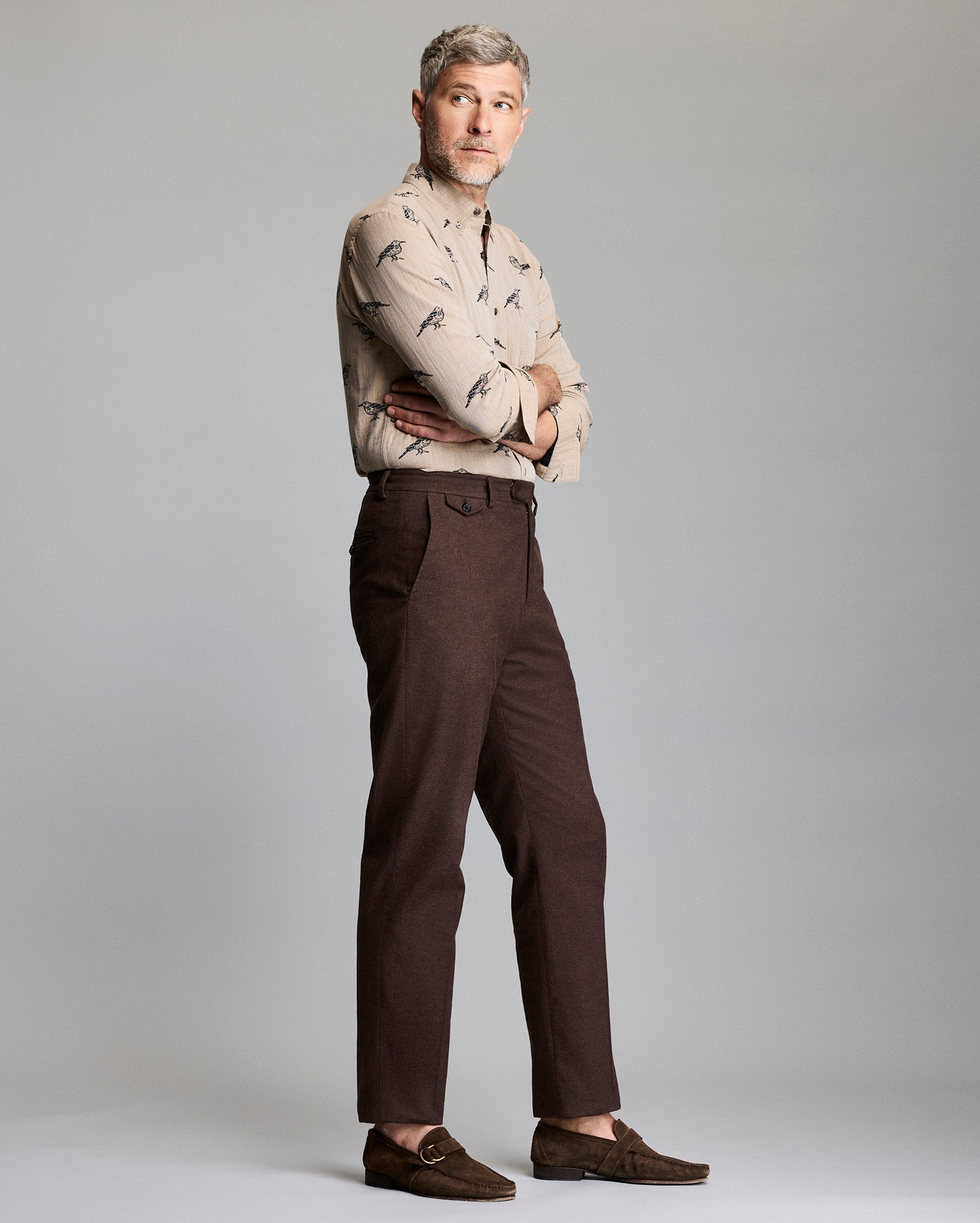 A man with short gray hair stands sideways with arms crossed, wearing a beige patterned button-up shirt, brown pants, and brown loafers, against a plain light gray background.