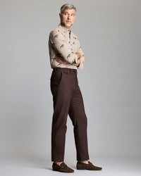 A man with short gray hair stands sideways with arms crossed, wearing a beige patterned button-up shirt, brown pants, and brown loafers, against a plain light gray background.