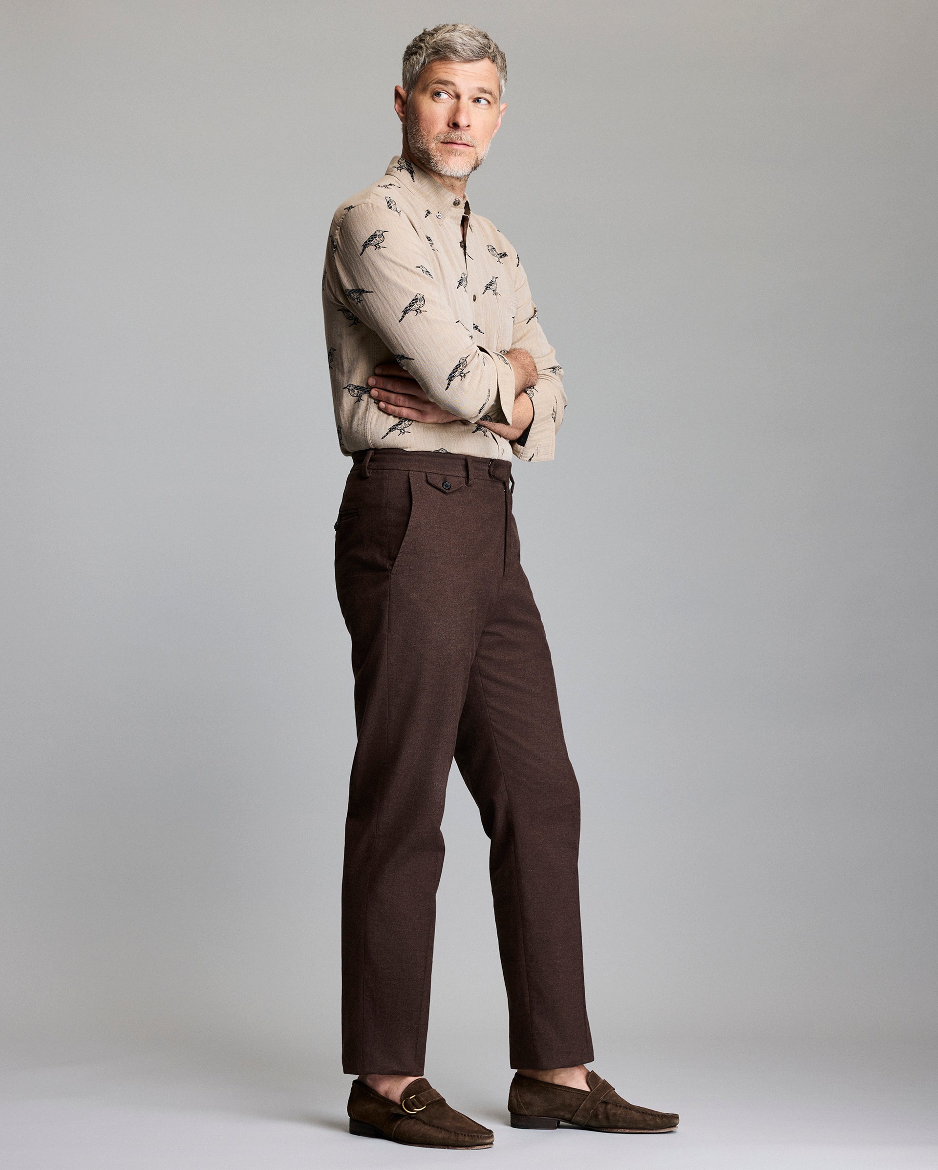 A man with short gray hair stands sideways with arms crossed, wearing a beige patterned button-up shirt, brown pants, and brown loafers, against a plain light gray background.