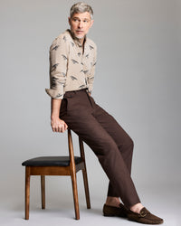 A man with short gray hair and a beard sits sideways on a wooden chair. He wears a tan patterned shirt, brown trousers, and brown loafers. The background is plain light gray.