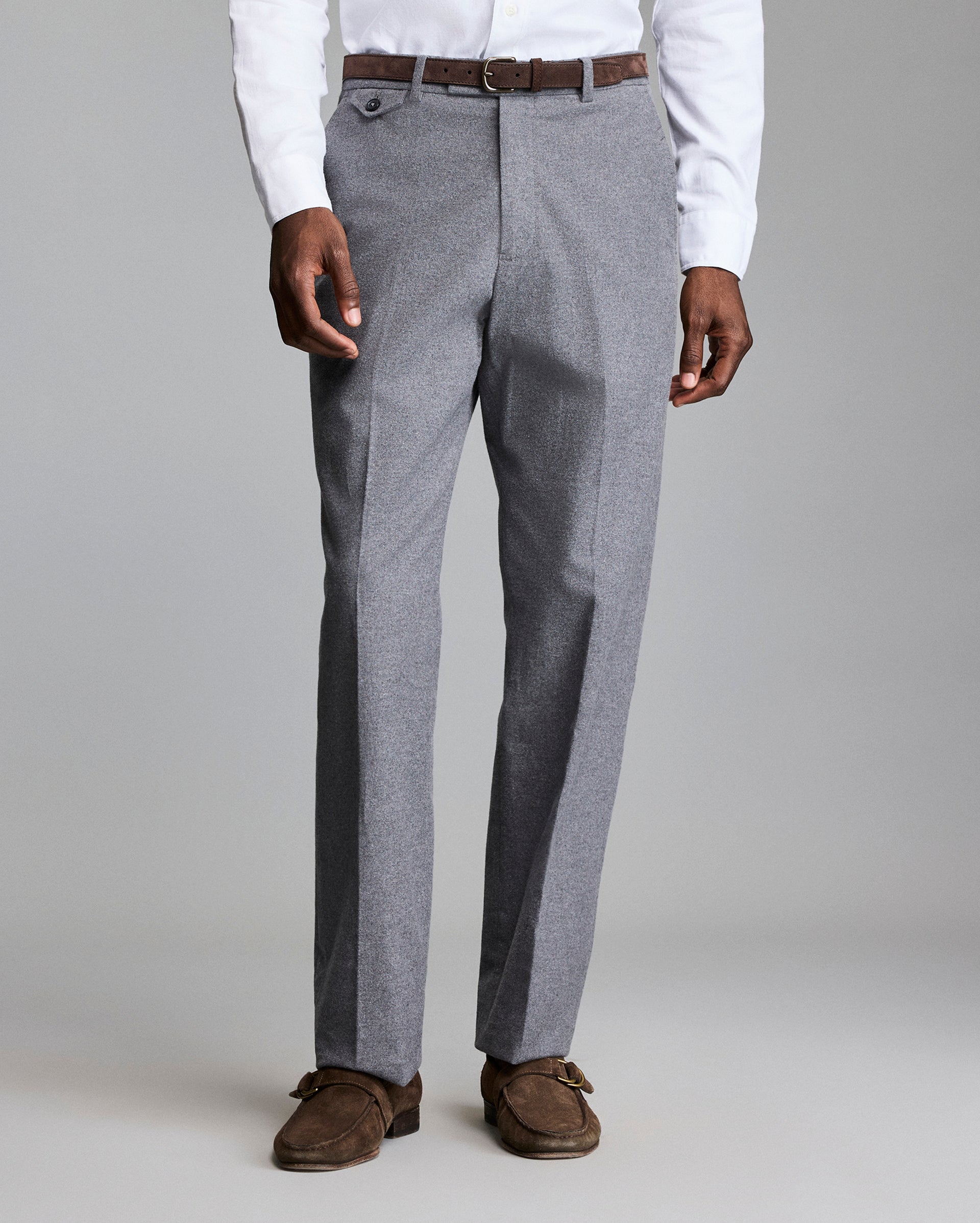 A person wearing gray dress pants, a white shirt, a brown belt, and brown suede loafers stands against a plain, light background. The image focuses on the lower half of the body.