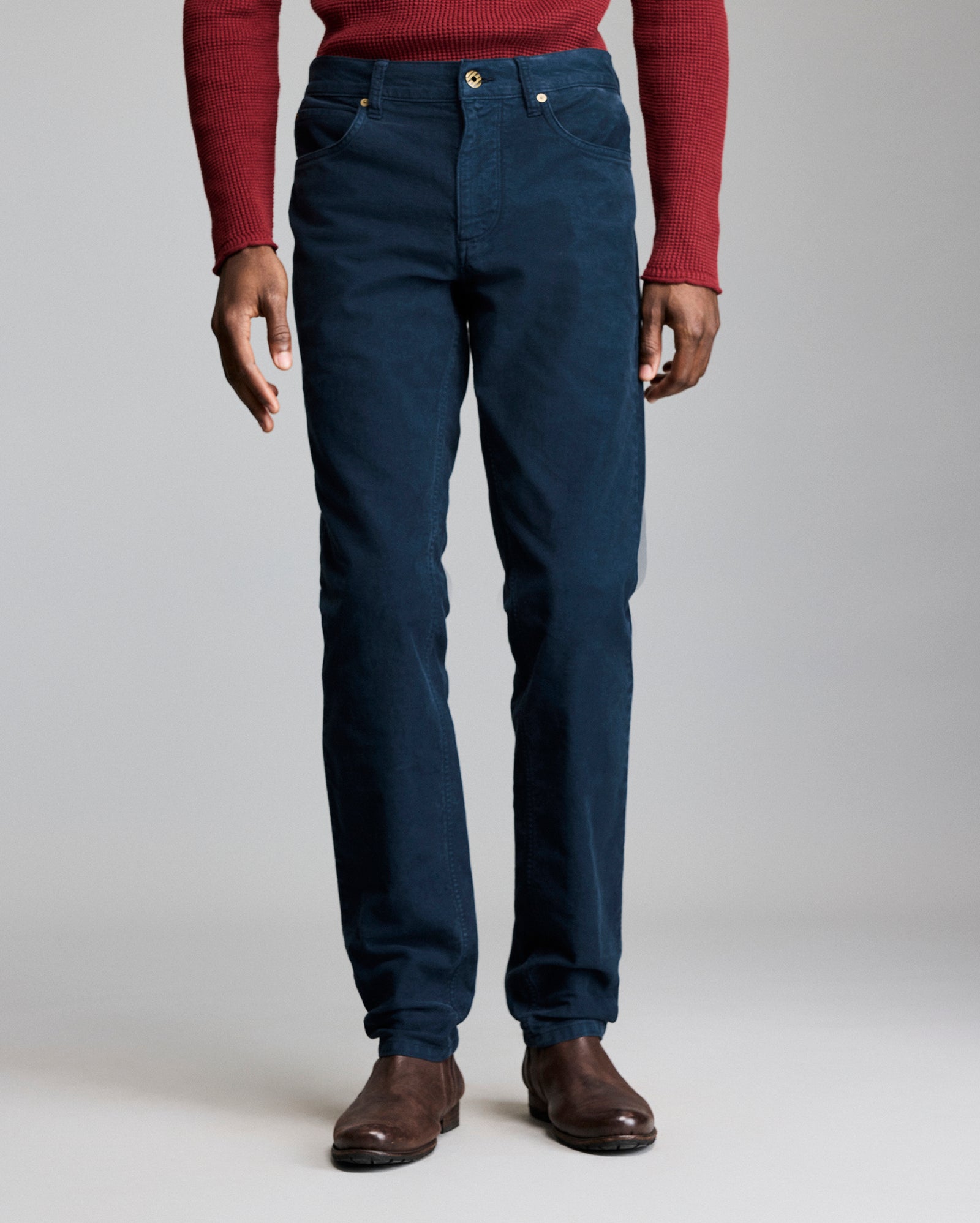 A person wearing a red long-sleeve shirt, dark blue pants, and brown shoes stands against a plain gray background, shown from shoulders to feet.