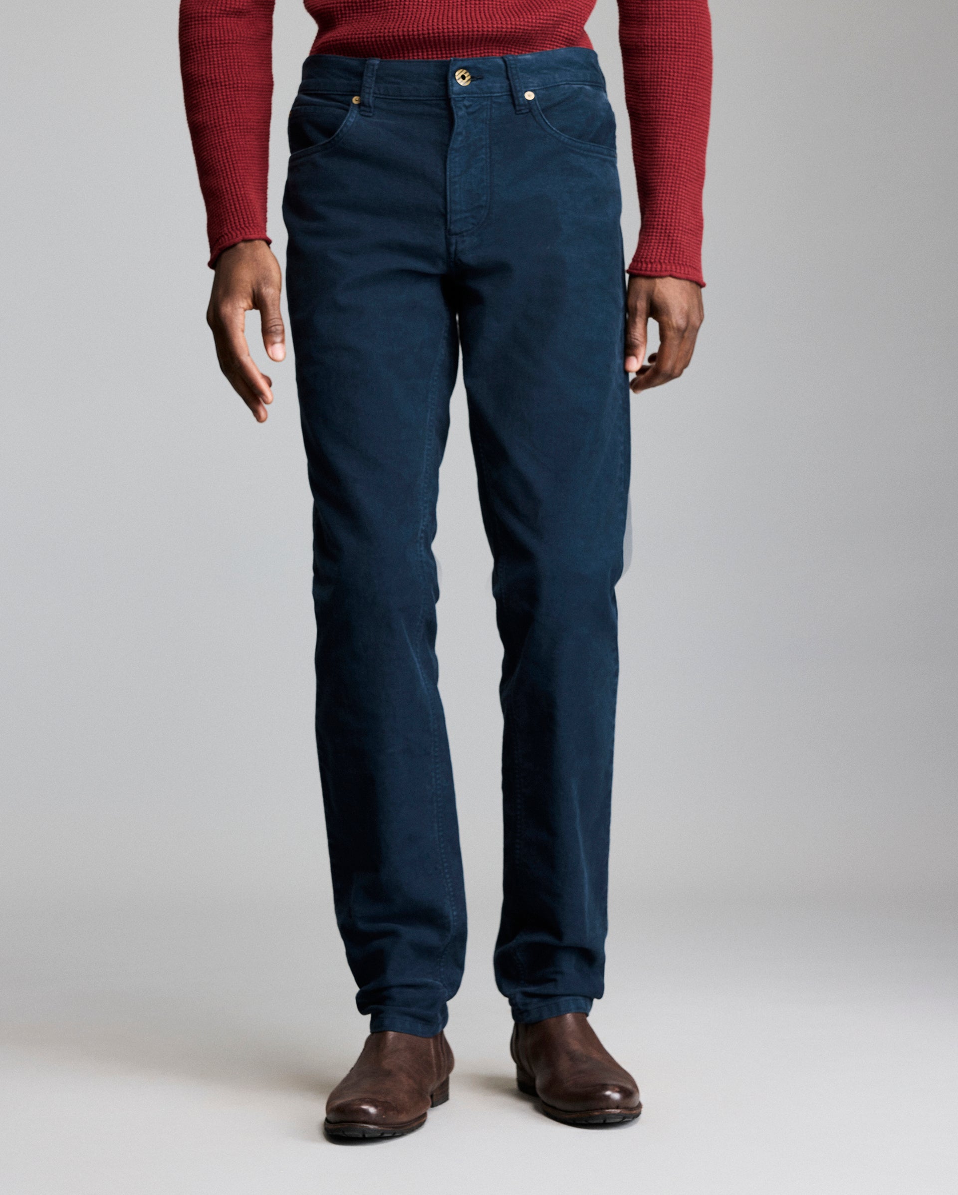 A person wearing a red long-sleeve shirt, dark blue pants, and brown shoes stands against a plain gray background, shown from shoulders to feet.