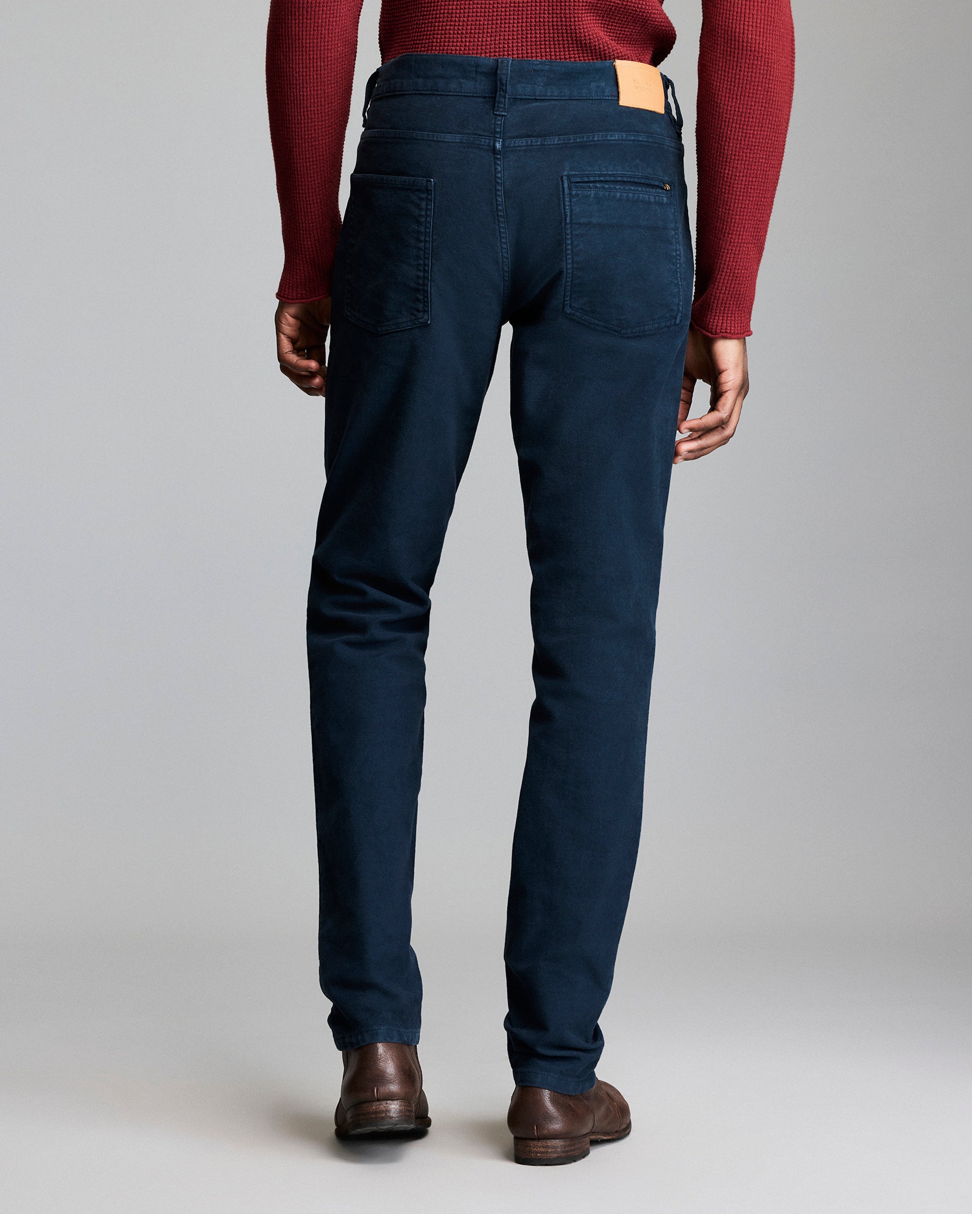 A person stands facing away, wearing a red sweater, dark blue pants, and brown shoes. The pants have two back pockets and a tan label on the waistband. The background is plain and light gray.