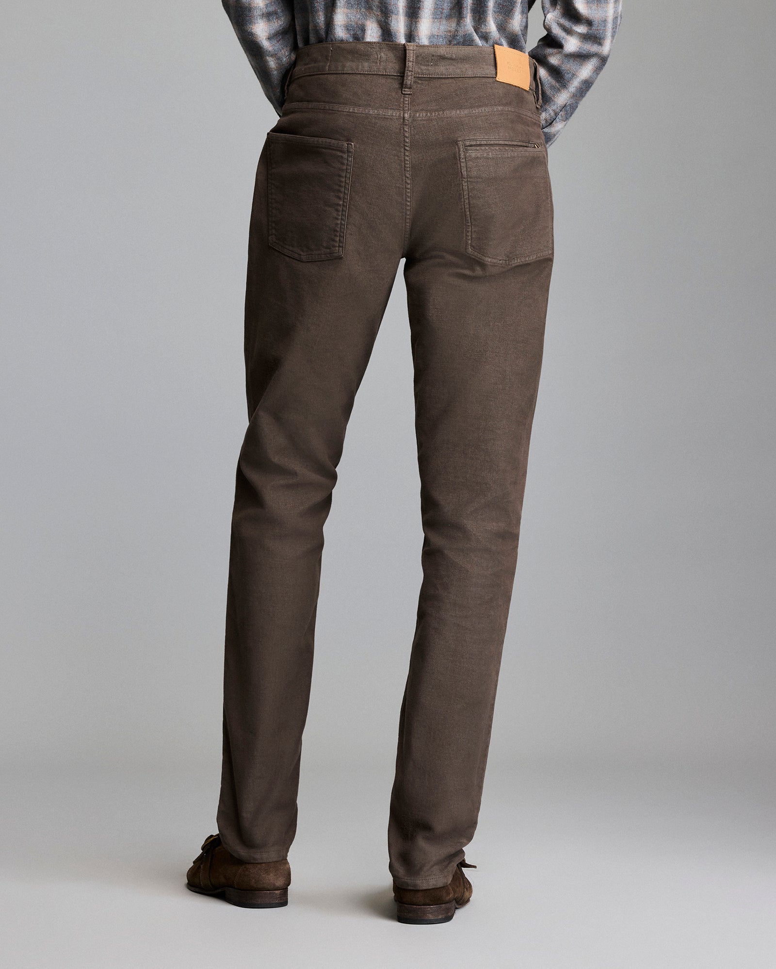 A person wearing brown pants, brown shoes, and a plaid shirt stands facing away from the camera against a plain gray background.